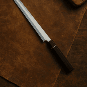 Yanagiba sashimi knife resting on worn brown leather with wood handle and single bevel blade, top-down view.